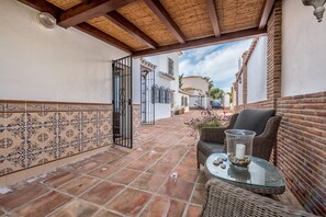Une terrasse couverte avec un sol carrelé, des meubles en osier et une table en verre agrémentée d’objets décoratifs.