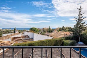 A view from a balcony overlooking a coastal area with terracotta-roof buildings, a swimming pool, and a clear blue sky.