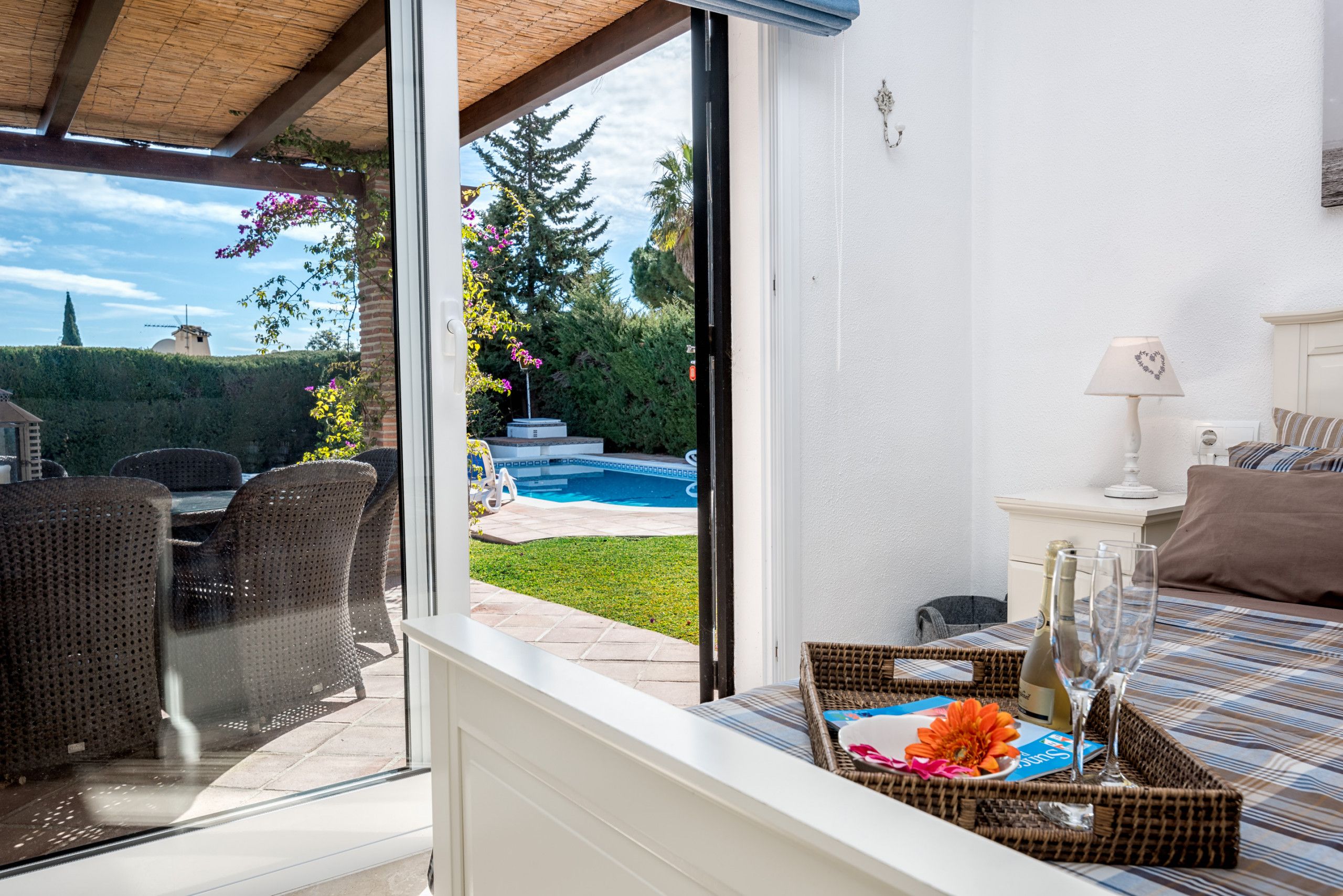 Una habitación con puerta de vidrio que da a una zona de piscina, muebles de mimbre, una mesita de noche con lámpara y una cama con colcha a rayas.
