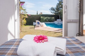 A bed with a white towel and a pink flower, a plaid blanket, and a view of a pool and garden outside.