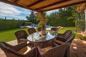 Eine überdachte Terrasse im Freien mit Korbmöbeln, einem Glastisch und einer Zierpfeiler.