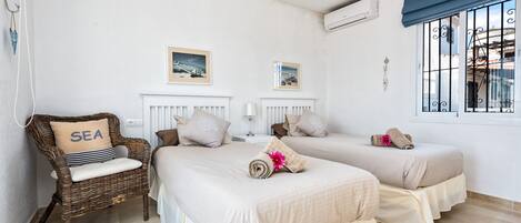 A hotel room with two beds, a wicker chair with a pillow labeled "SEA", and a window with bars.