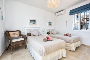 A hotel room with two beds, a wicker chair with a pillow labeled "SEA", and a window with bars.