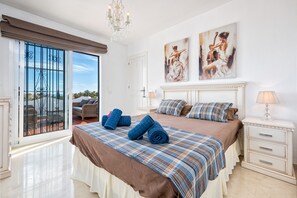 A bedroom with a large bed, a nightstand, a chandelier, and a view through a glass door.