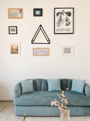 Ein Zimmer mit einem blauen Sofa, einem Bilderrahmen mit einer Kaktee-Darstellung und einem Bilderrahmen mit einem Frauenporträt.