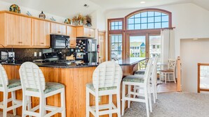 A kitchen with wooden cabinets, a black refrigerator, and a bar area with white chairs. - Love R Shack: Oceanside Duplex, Spa, Shared Pool (Corolla)