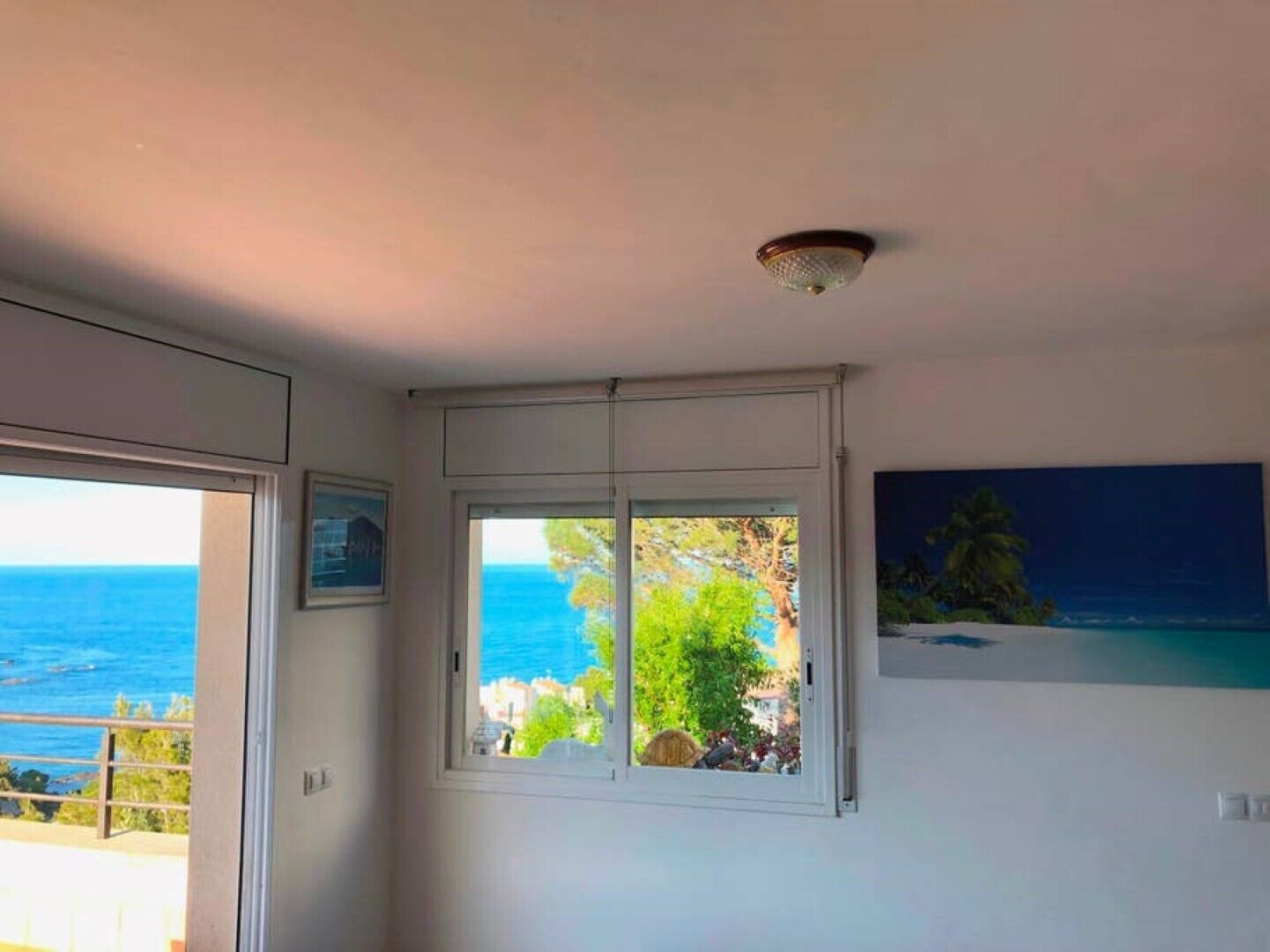 Quarto com uma grande janela que oferece vista para o mar e vegetação, um lustre e um quadro na parede.