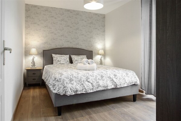 A neatly made bed with a floral bedspread, two bedside lamps, a nightstand, and a wooden floor.