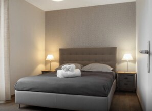 Una cama bien hecha con un cabecero gris, dos mesitas de noche con lámparas y un pequeño aparador.