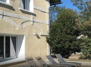 Un edificio con fachada color beige, marcos de ventana blancos y una pequeña lámpara blanca. Hay sillones reclinables alineados contra la pared del edificio.