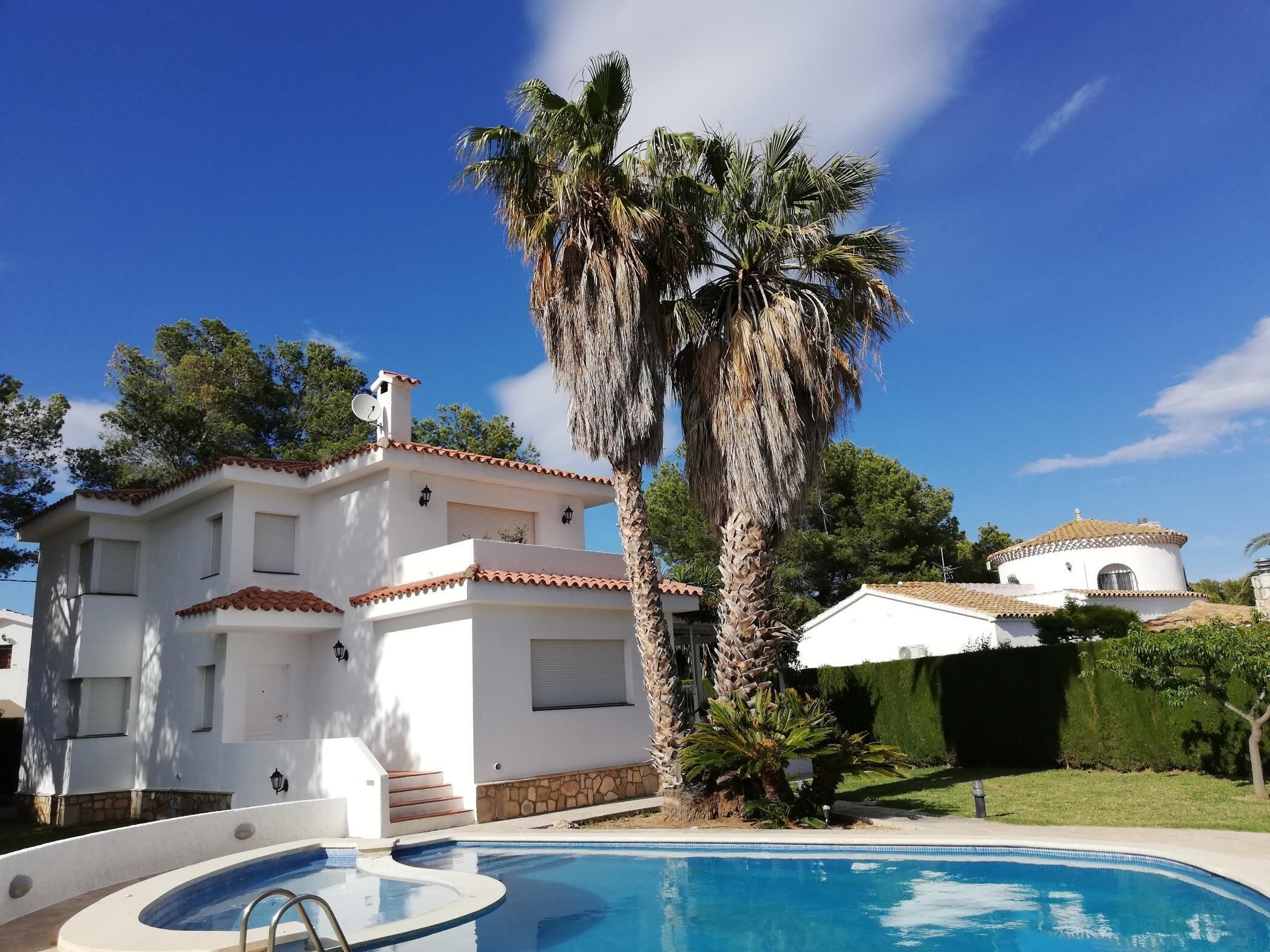 Une maison blanche à deux étages, dotée d’une piscine, de palmiers et d’un petit jardin.