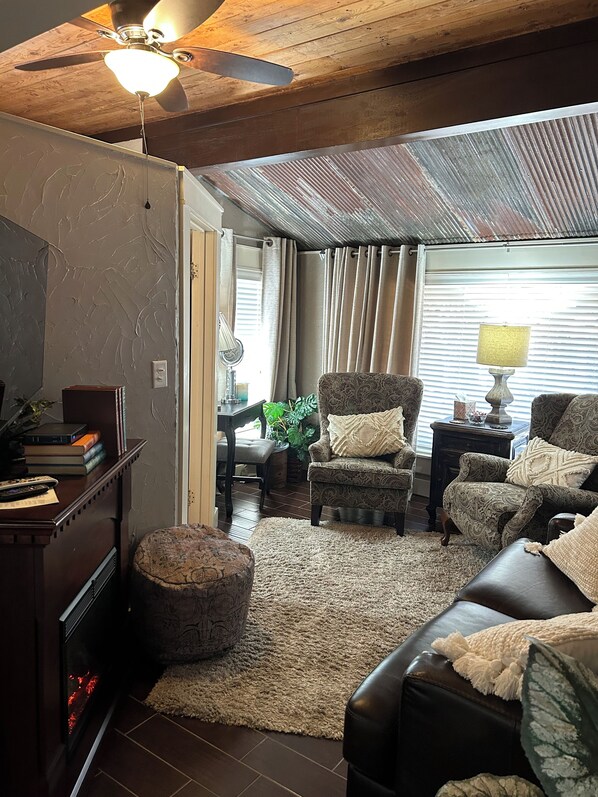 A cozy living room with a fireplace, a ceiling fan, and a large window with curtains.