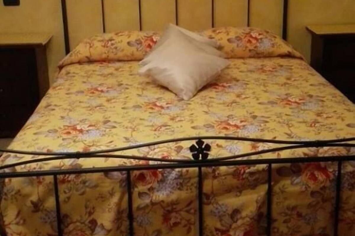 Un lit avec une couvre-lit à motifs floraux et un seul oreiller, une lampe fixée au mur et un tableau encadré au-dessus du lit.