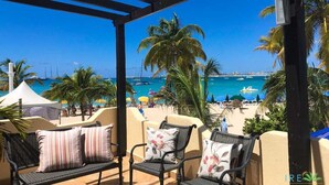 A covered outdoor seating area with wicker chairs and cushions, overlooking a beach with palm trees and boats.