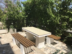 Un espace extérieur aménagé en patio, avec une table et des bancs, entouré de verdure et doté d’un sol en pierre.
