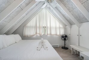 Une chambre avec un grand lit, un ventilateur sur pied et une fenêtre ornée de rideaux légers.