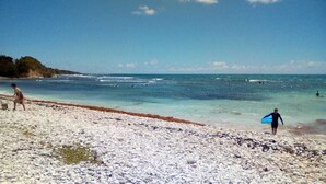 A beach with clear water, a rocky foreground, and a person with a surfboard. - Villa Ti Colibri in Sainte-Anne 350 m from the sea (Sainte-Anne)