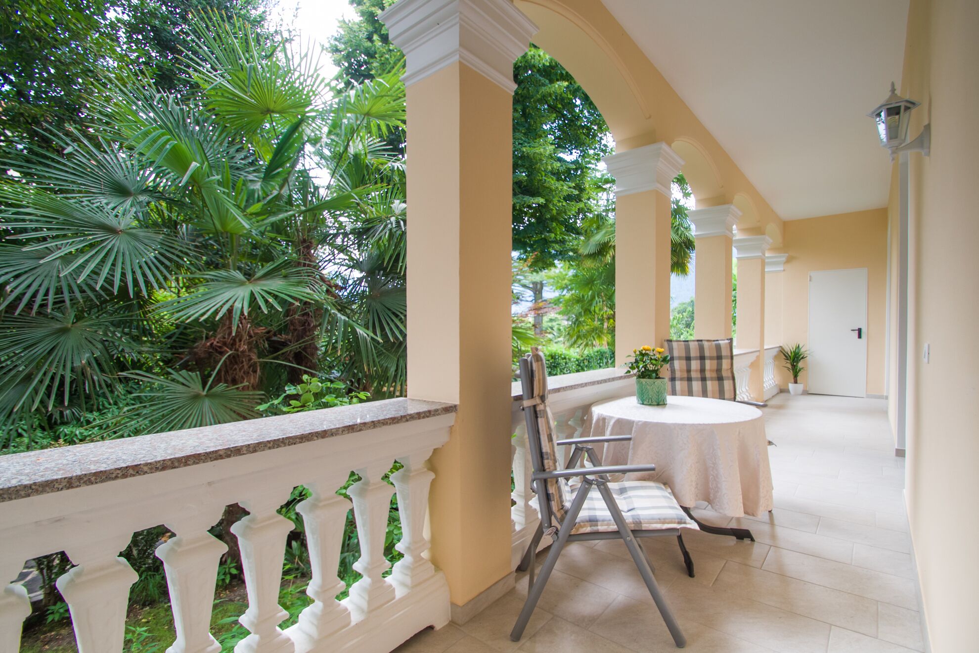A covered outdoor seating area with a table and chairs, surrounded by lush greenery and a white balustrade.