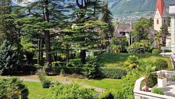 A lush garden with mature trees, a pathway, and a church with a red roof. - Residence Diana (Merano)