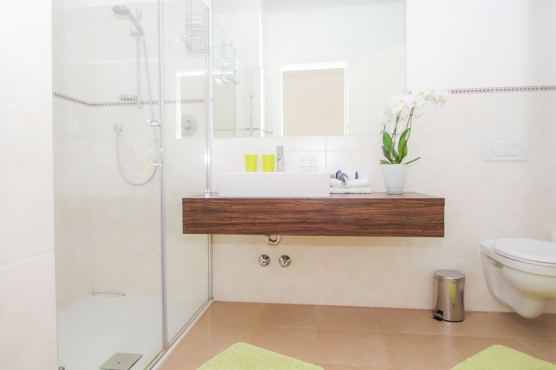 A modern bathroom with a glass shower enclosure, a wooden vanity with a mirror, a toilet, and a trash bin.