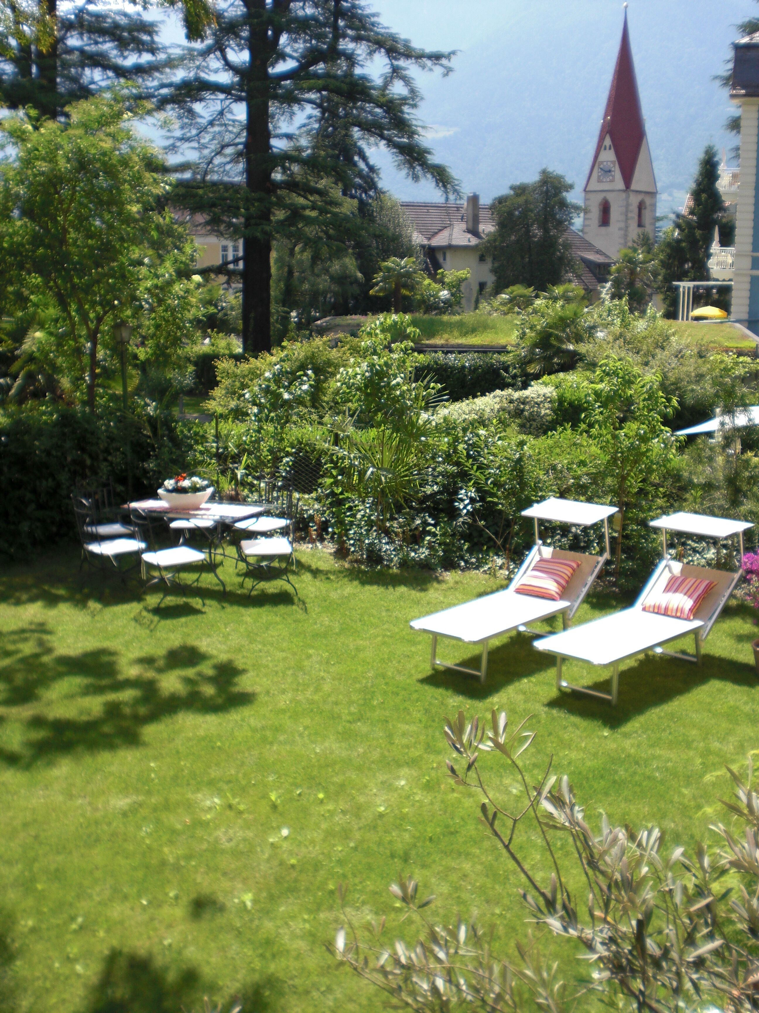 A garden with lounge chairs and tables, surrounded by greenery and a building with a red roof in the background.
