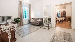 A spacious living room with a grey sofa, a glass coffee table, and patterned rugs. A chandelier hangs from the ceiling, and a large window provides natural light. - Residence Diana (Merano)