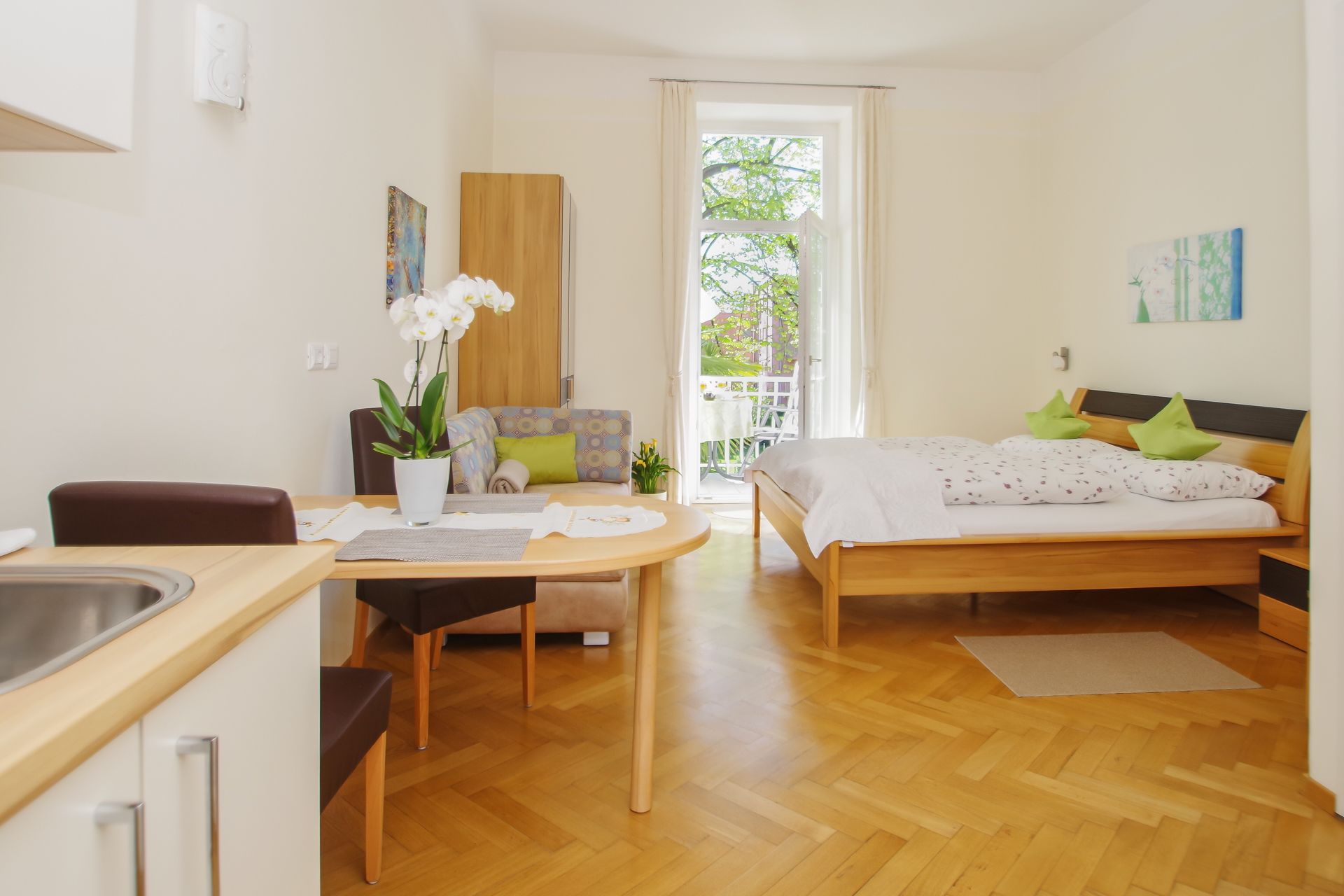 A modern bedroom with a wooden bed, a small desk, and a chair. There is a balcony with a view of greenery.