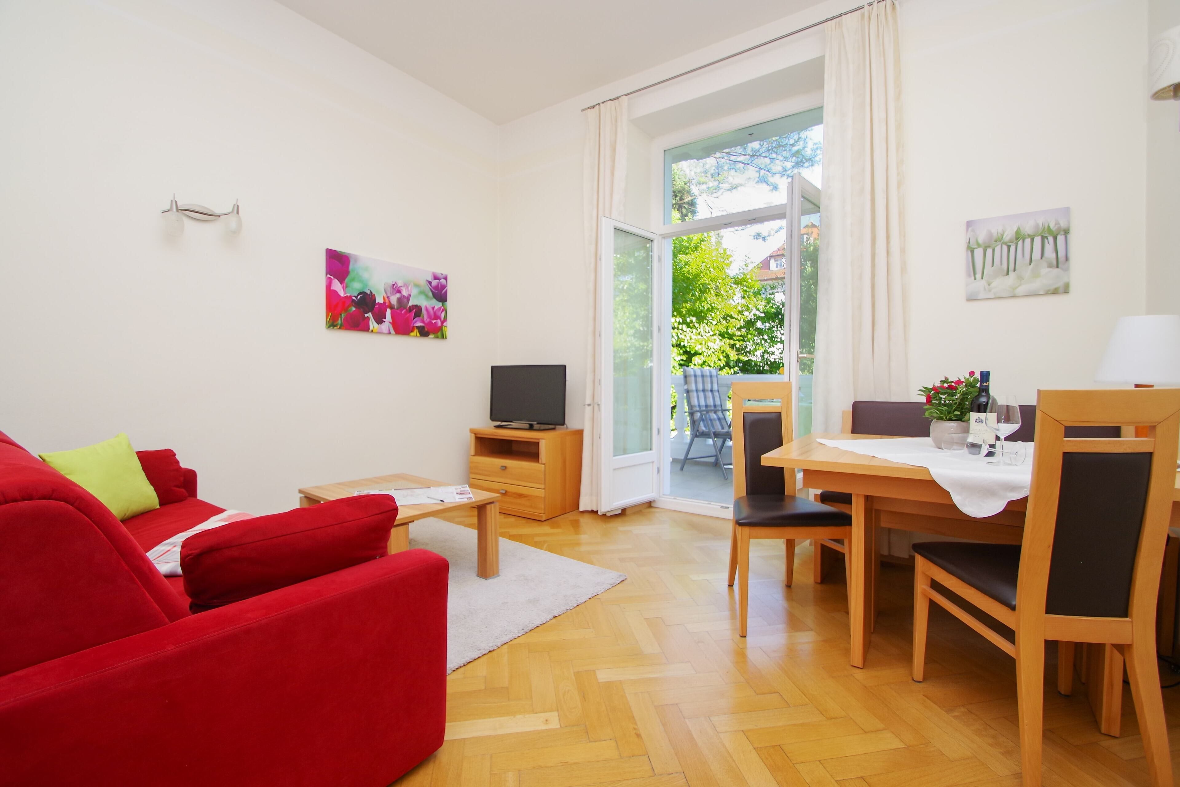 A living room with a red sofa, a TV, a dining table with chairs, and a sliding glass door leading to an outdoor area.