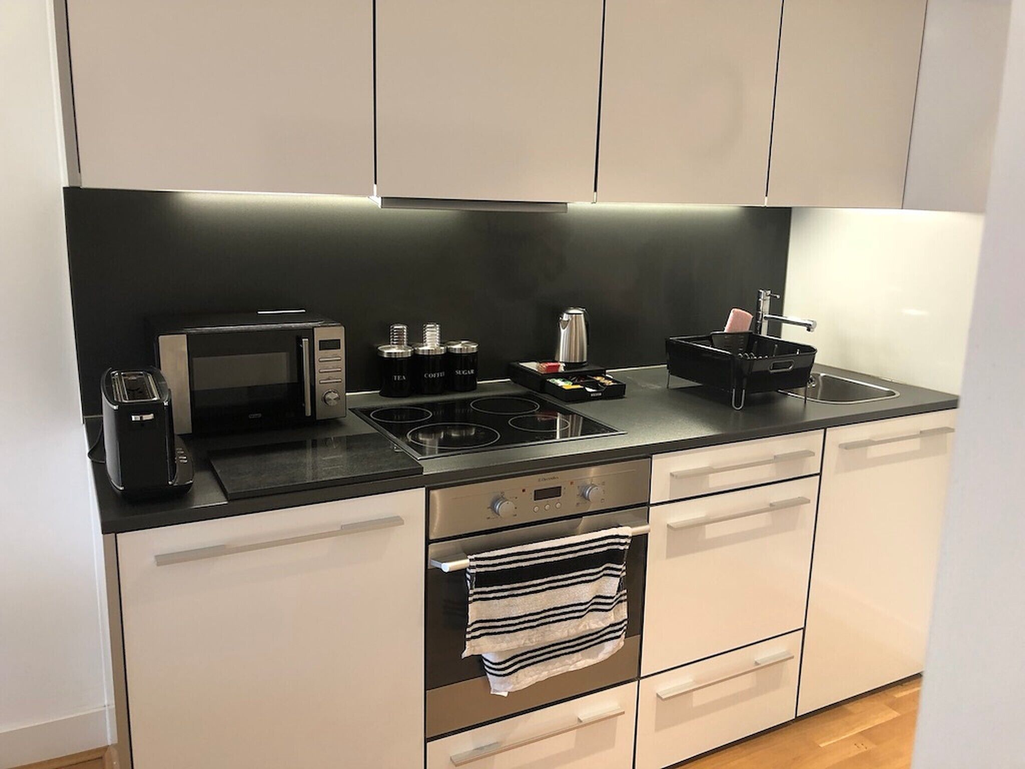 A modern kitchen with a black countertop, a built-in oven, a microwave, and a kettle. There are cabinets above the countertop and a towel hanging from the oven handle.