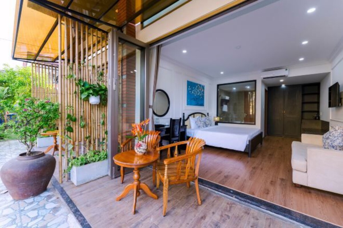 A modern living room with a sofa, a dining table with chairs, a large potted plant, and a bed in the background.