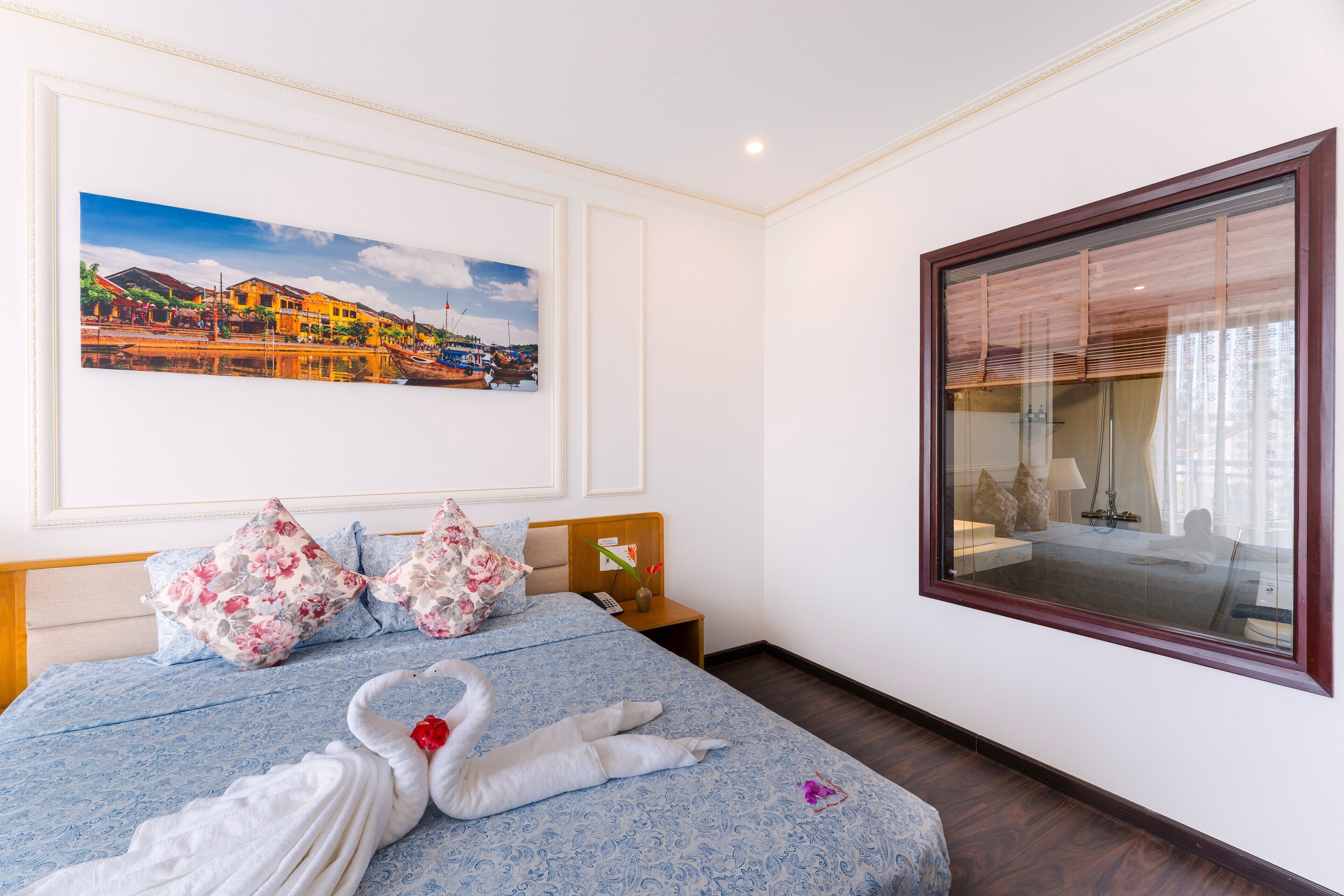 Habitación de hotel con cama, mesita de noche, un cuadro enmarcado y un ventanal grande.