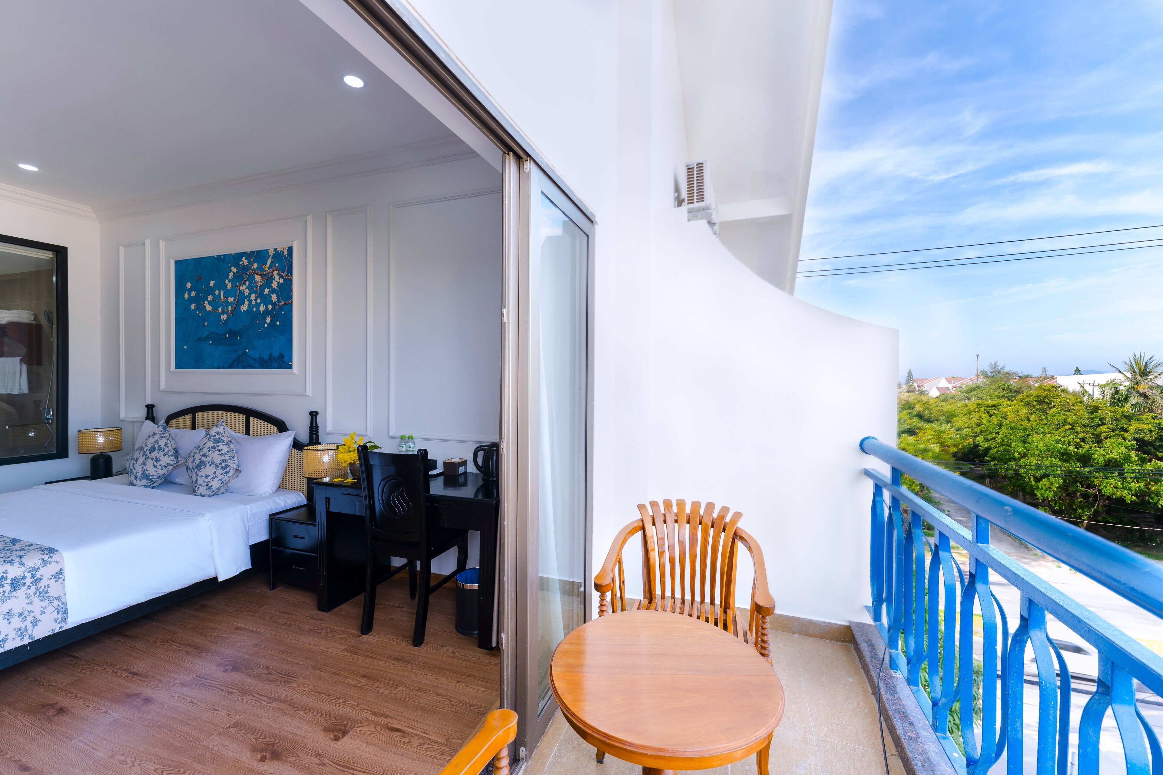A bedroom with a bed, a desk, and a chair, a balcony with a blue railing, and a painting on the wall.