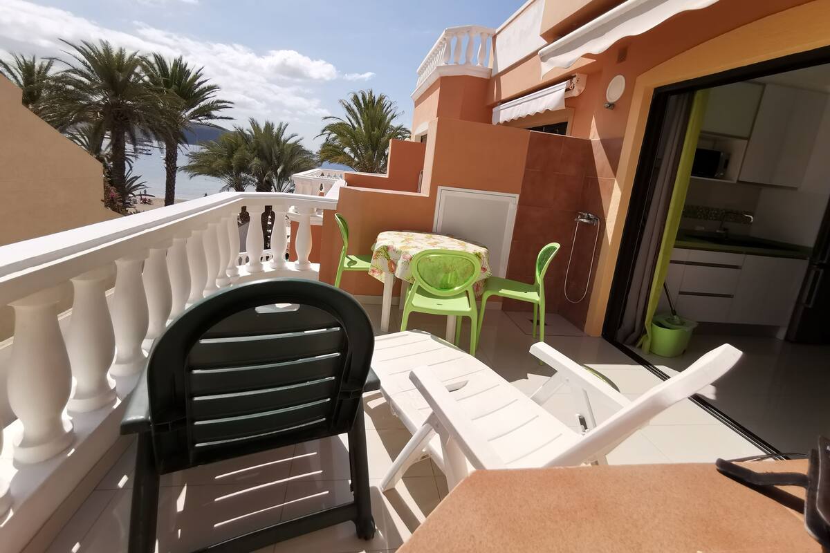 Un balcon doté d’une balustrade blanche, une table avec des chaises, et une vue sur des palmiers et un bâtiment en arrière-plan.