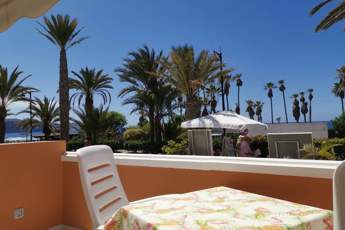 Un balcon avec une table recouverte d’une nappe à motifs de fruits, donnant sur une plage bordée de palmiers et surplombant un ciel bleu azur.