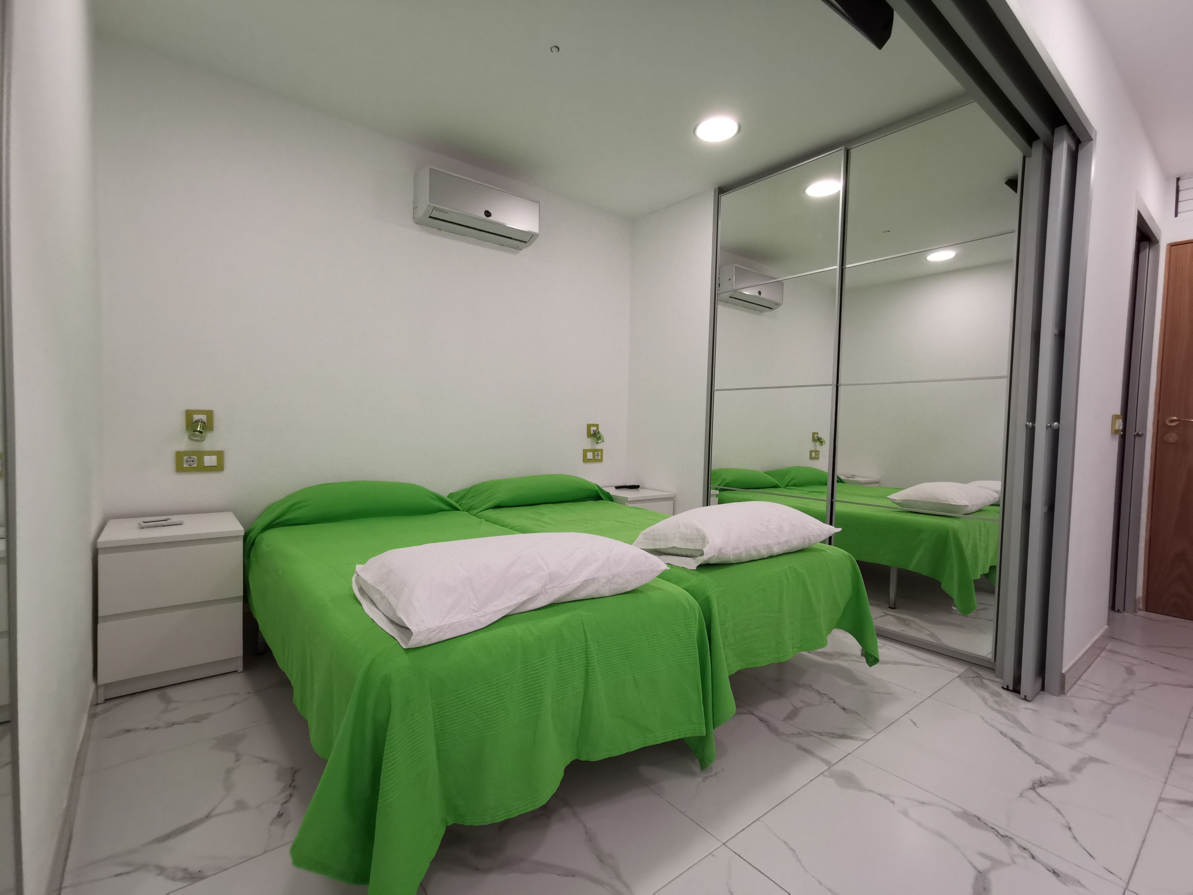 A hotel room with two beds, green bedding, a white nightstand, a mirror, and a marble-tiled floor.