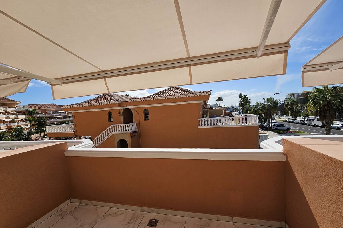 Un balcon avec un sol carrelé, une vue sur un immeuble avec un toit en tuiles, et une rue où l’on aperçoit des voitures et des palmiers au loin.