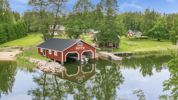 A red building labeled "PETTU" with a dock extending into a calm lake, surrounded by lush greenery and other houses. - Lilla nennebo by Interhome (Salo)