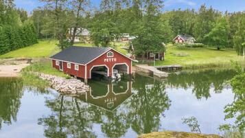 Un bâtiment rouge avec un garage portant l’inscription « PETTU », situé au bord d’un lac paisible aux eaux claires et aux reflets nets.
