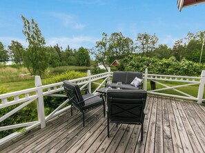 A wooden deck with a table and two chairs, surrounded by a white fence, overlooking a green landscape with trees and a lake. - Lilla nennebo by Interhome (Salo)