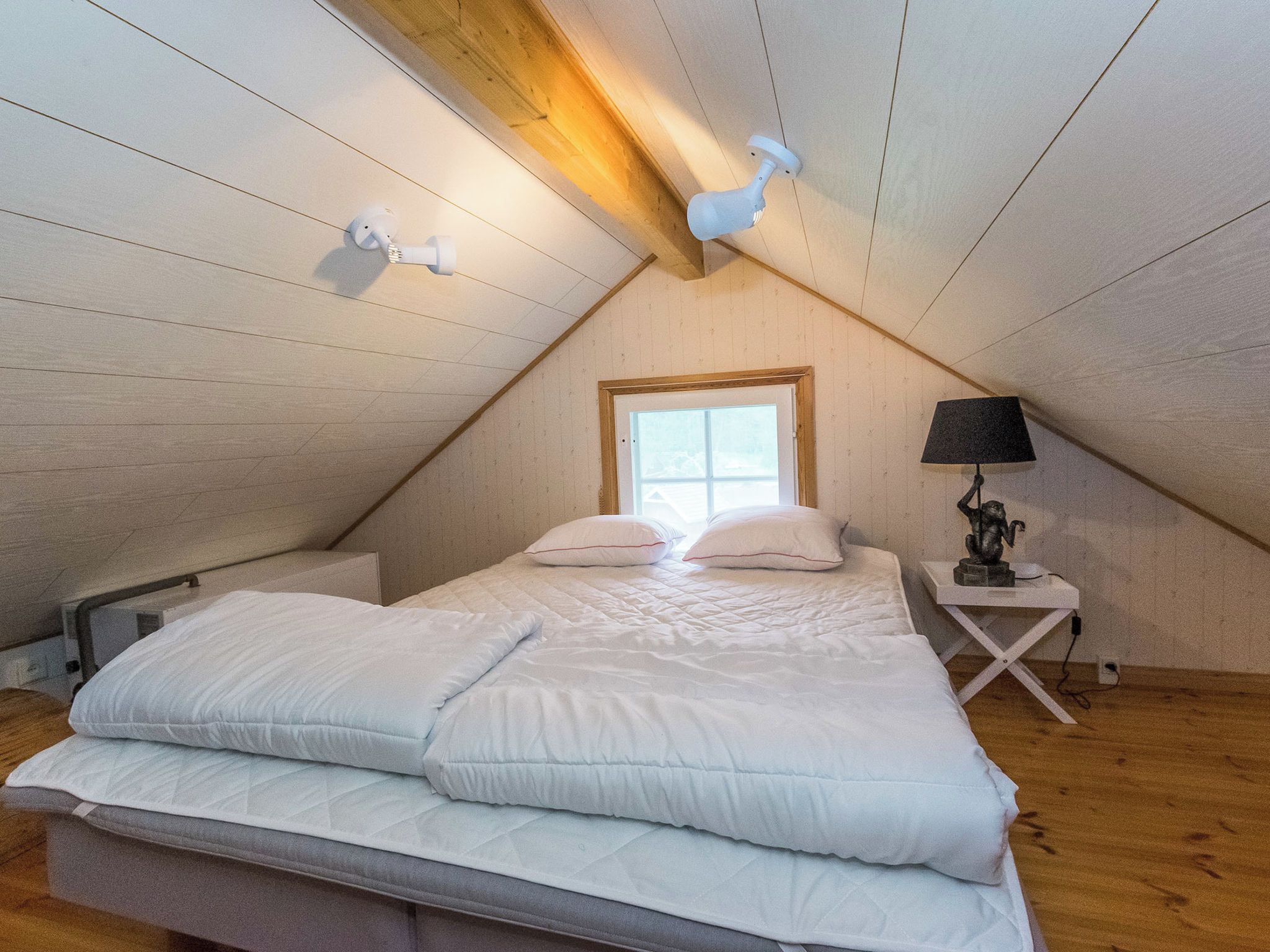 Et soveværelse med skrå vægge, en seng med hvide sengetøj, et vindue og en sengebordlampe.