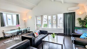 Una sala moderna con un sofá de cuero negro, una mesa de centro de vidrio y un comedor con mesa y sillas de vidrio. Amplias ventanas con cortinas dejan entrar luz natural y ofrecen vistas de la ciudad.
