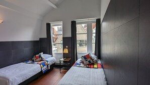 Una habitación pequeña y moderna con dos camas, una mesita de noche y una lámpara. Cuenta con ventanas grandes con cortinas y suelo de baldosas.
