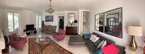 Uma sala de estar ampla, com uma televisão grande, assentos confortáveis, um lustre e vista para o exterior.
