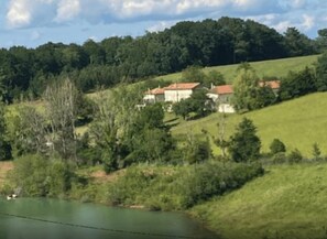 A rural landscape with a body of water, a hill with trees, and a few buildings with red roofs.
