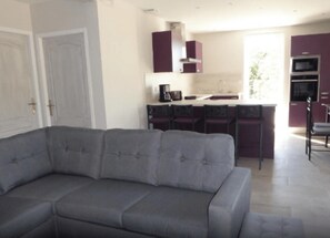 A modern living room with a grey sofa, a kitchen area with purple cabinets, and a dining area with a table and chairs.