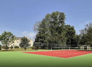 Un court de tennis avec un filet, entouré d’arbres et avec un bâtiment en arrière-plan.