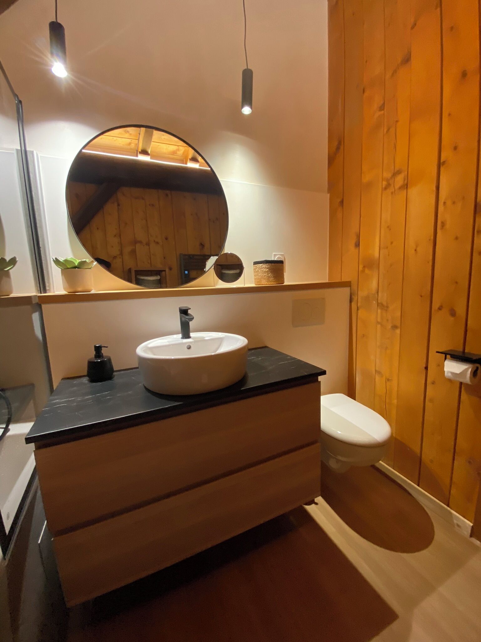 Een badkamer met een ronde spiegel, een witte wastafel, een zwart aanrecht, een houten wastafelmeubel en een toilet.