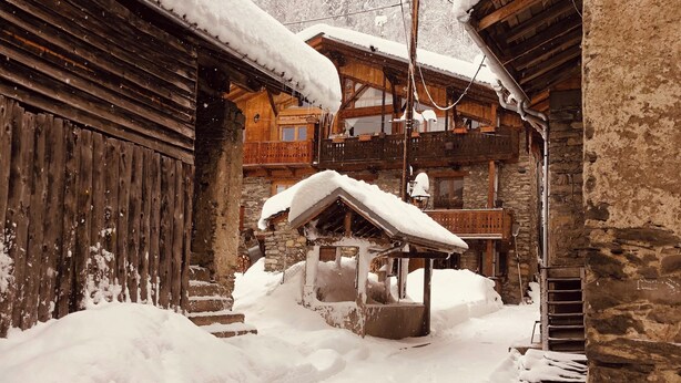 A snow-covered wooden building with a stone wall and a wooden staircase leading to a second-floor balcony.