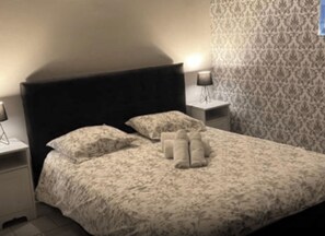 A neatly made bed with a floral bedspread, two pillows, and two rolled towels. There are two bedside tables with lamps, one on each side of the bed. - GITE COSMOS (Notre-Dame-de-Sanilhac)