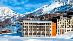 Un hôtel à plusieurs étages, une piste de ski en arrière-plan et des montagnes enneigées.